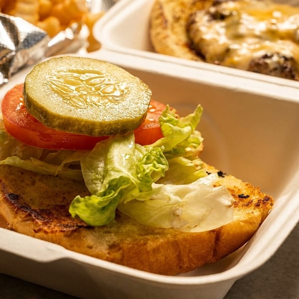 Open-faced burger with lettuce, tomato, and pickle in a takeout container, with another burger behind it.