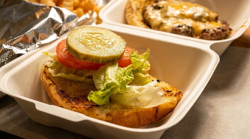 Open-faced burger with lettuce, tomato, and pickle in a takeout container, with another burger behind it.