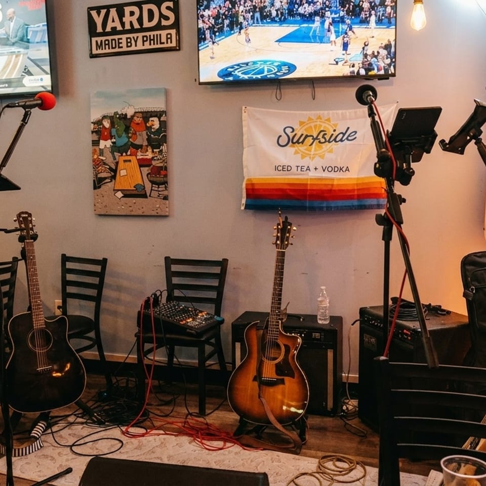 A bar interior with guitars, microphones, speakers, TVs, and a dartboard, set up for live music.