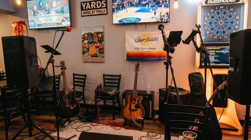A bar interior with guitars, microphones, speakers, TVs, and a dartboard, set up for live music.