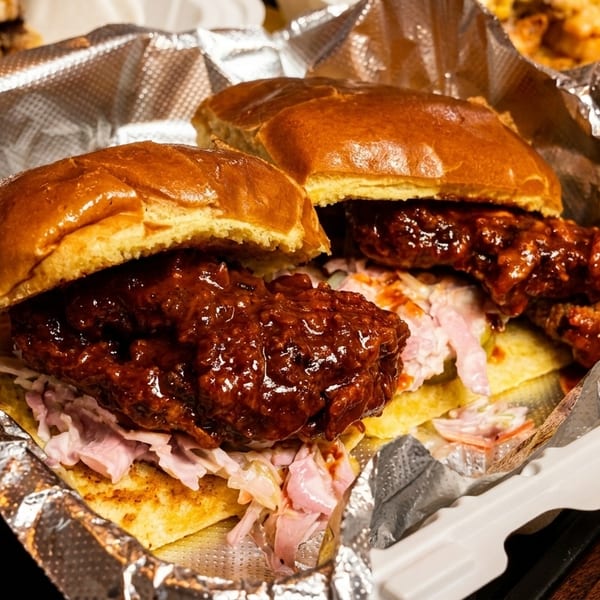 Two saucy chicken sandwiches with coleslaw on brioche buns in a foil-lined takeout container.