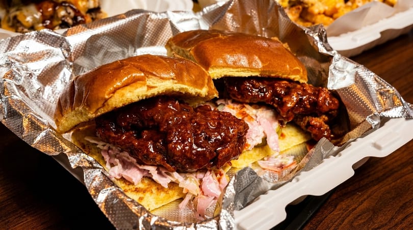 Two saucy chicken sandwiches with coleslaw on brioche buns in a foil-lined takeout container.
