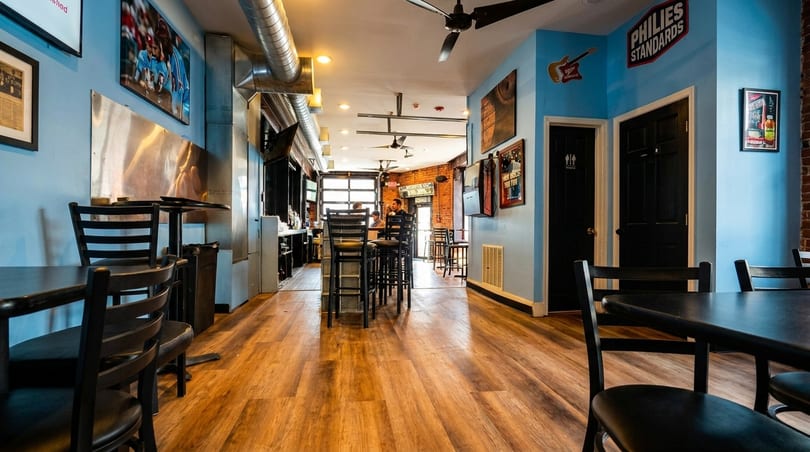 Interior of a restaurant with light blue walls, wooden floors, black tables, and a bar area.