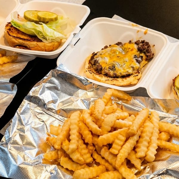 Two cheeseburgers in takeout containers with crinkle-cut fries on foil, ready to eat.