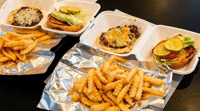 Two cheeseburgers in takeout containers with crinkle-cut fries on foil, ready to eat.
