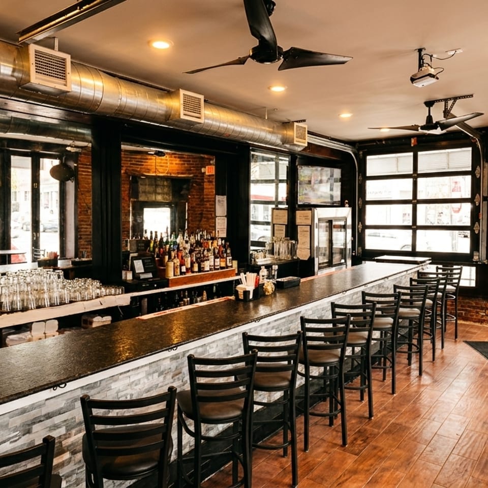 A long bar with stools and tables in a restaurant with exposed brick walls and large windows.