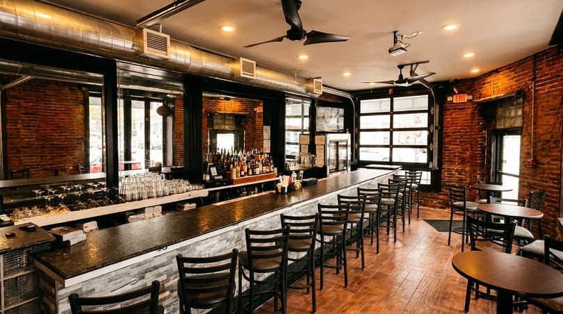 A long bar with stools and tables in a restaurant with exposed brick walls and large windows.