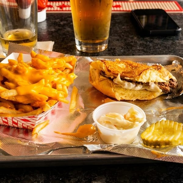 A tray with a cheesesteak, cheese fries, coleslaw, and a pickle, with a beer in the background.