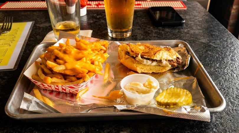 A tray with a cheesesteak, cheese fries, coleslaw, and a pickle, with a beer in the background.